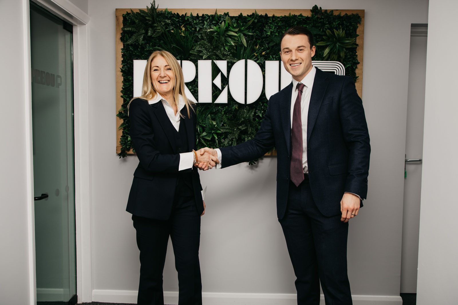 Two members of the Preou team shaking hands in front of the Preou logo on a planted wall